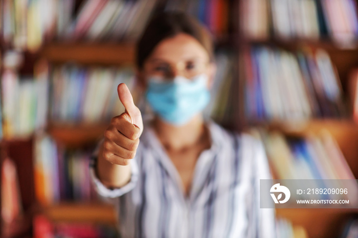 Closeup of college girl standing in library and holding thumbs up. She is having face mask to preven