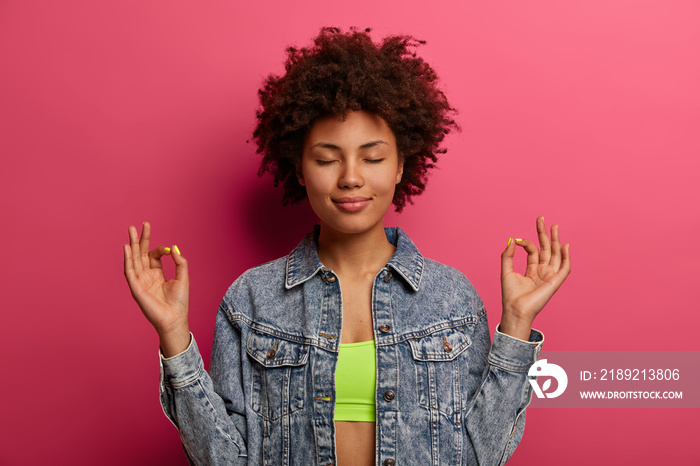 Portrait of relaxed satisfied woman makes yoga gesture, meditates with eyes closed, dressed in fashi