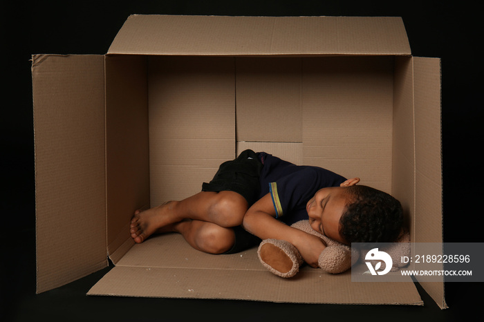 Cute little boy living in box on dark background. Poverty concept