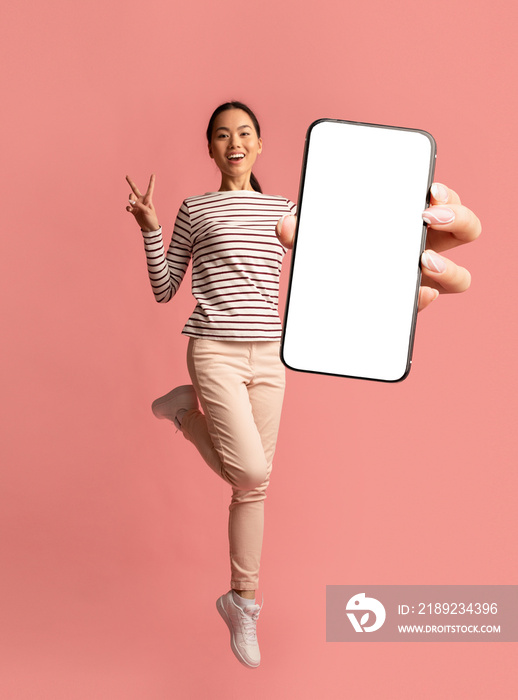 快乐的千禧一代亚洲女性拿着智能手机跳跃，在粉色工作室背景上展示和平姿态