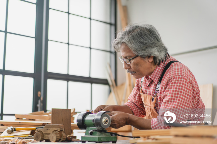 Asian senior carpenter man using small grinder machine make wooden car at workshop