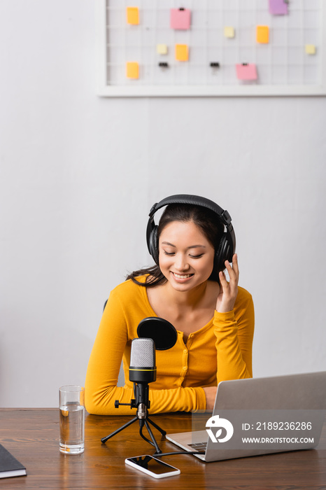 young asian radio host touching wireless headphones while sitting near microphone, laptop and smartp