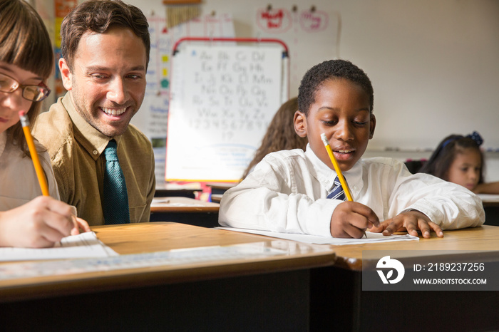 Portrait of male teacher looking at schoolgirl (10-11) and schoolboy (8-9) writing at desks in class