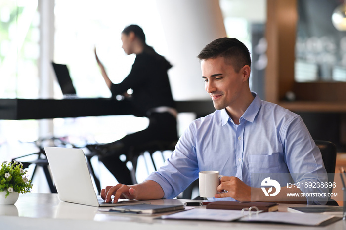 Handsome businessman holding coffee cup and using laptop computer in modern office.