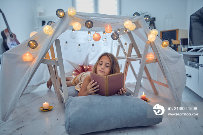 Little 10 year old girl reading classic book under her home-made tent inside the living room.