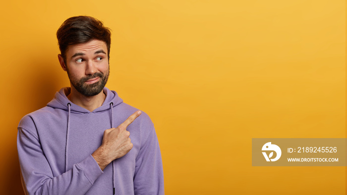 Horizontal studio shot of unimpressed bearded man indicates at blank space, has curious face express