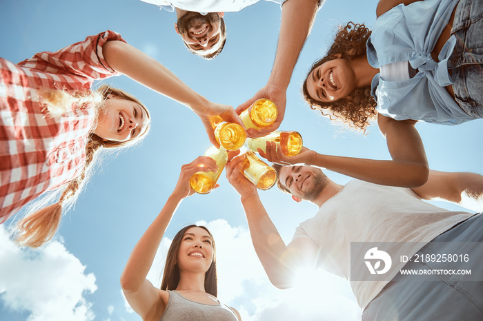 Enjoying summer time. Happy young people are clinking bottles with beer and smiling against blue sky