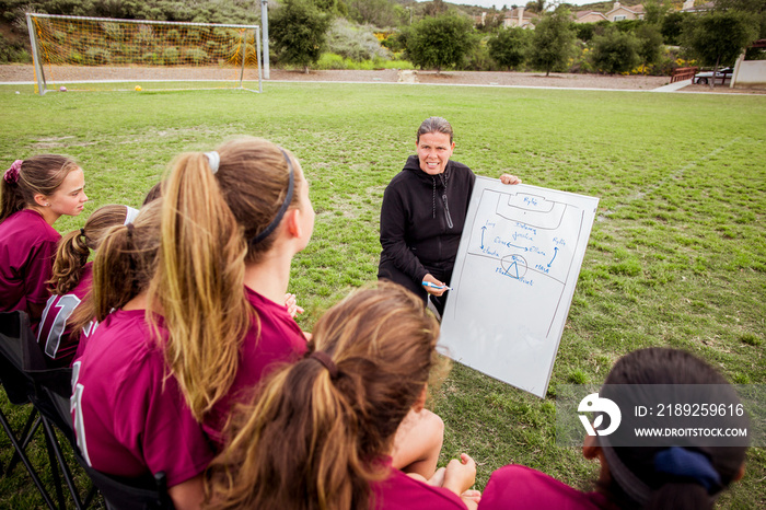 Girls in sports jersey looking at coach making plan over whiteboard during competition on field