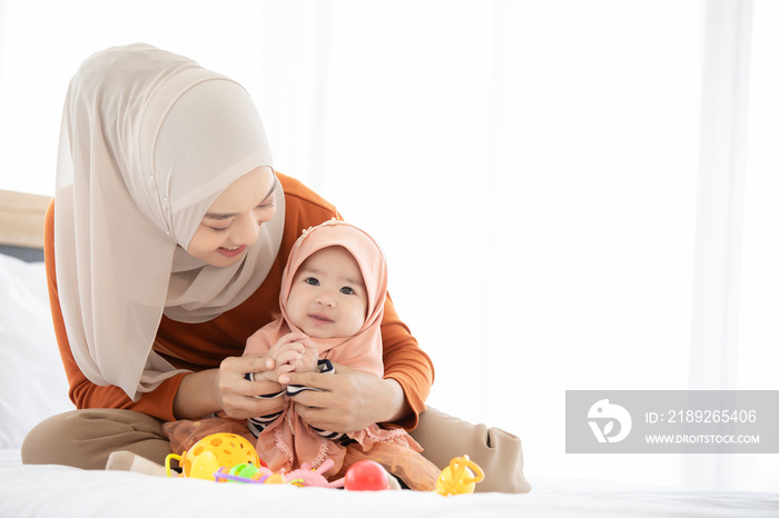A Muslim mother and  toddler are indoors in their bedroom. The mother is wearing a head scarf, and s