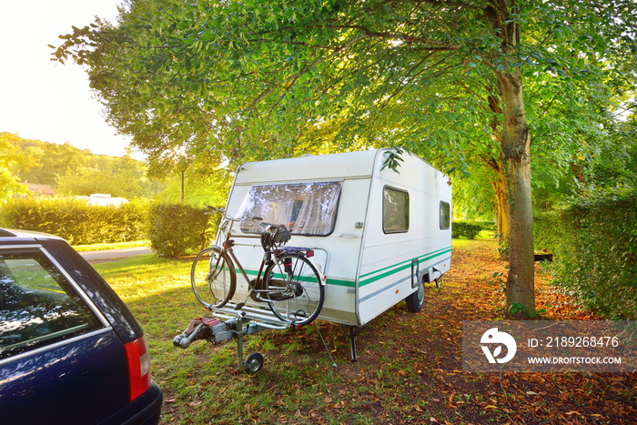 Caravan trailer with a bicycle and a car parked on a green lawn under the trees in a camping site. N