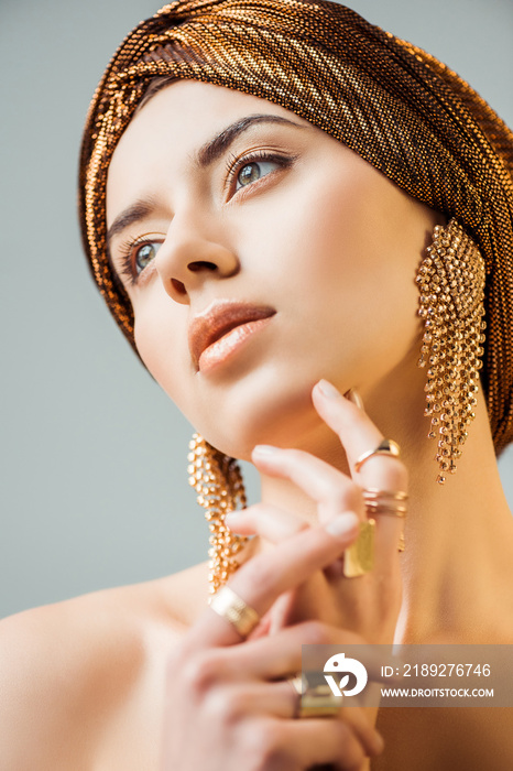 young naked woman with shiny makeup, golden rings and earrings in turban isolated on grey