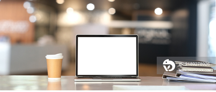 White blank screen laptop, coffee cup and document folder on the working desk over blurred office ro