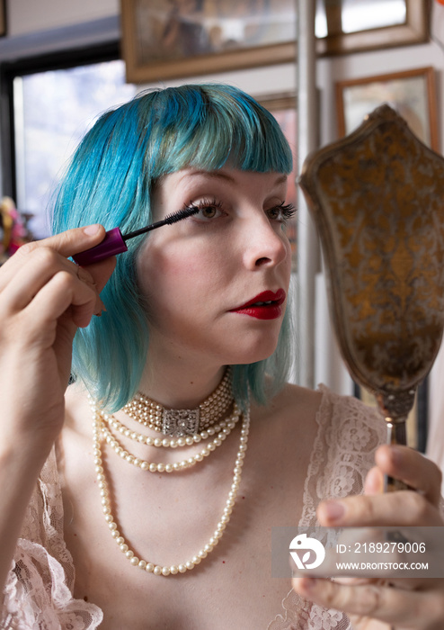 woman with blue hair applying mascara with antique mirror