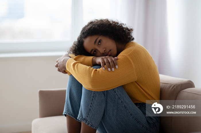 Young depressed black woman hugging her knees on sofa at home, absorbed in negative thoughts, having