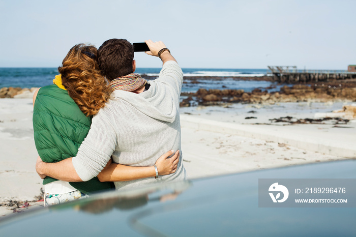 Rear view of young couple leaning against car taking selfie on smartphone, Cape Town, Western Cape, 