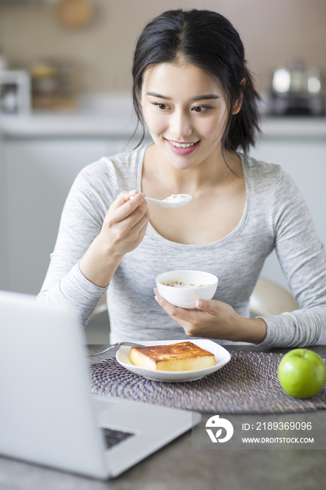 年轻女子一边吃早餐一边使用笔记本电脑