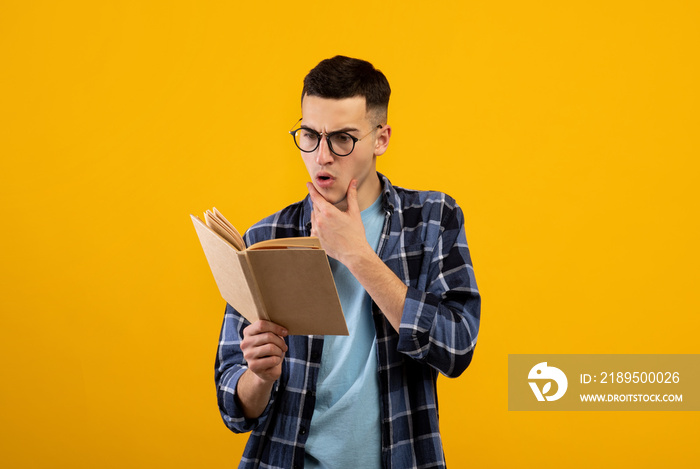 Shocked young student in glasses reading textbook, having difficulty studying for exam on orange stu