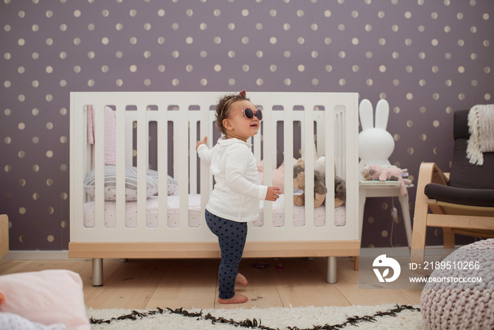 Side view of cute baby girl wearing sunglasses standing by crib against wall at home