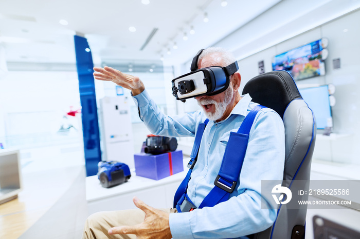 Excited bearded senior man sitting in chair and trying out virtual reality goggles. Tech store inter