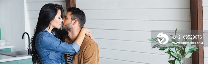 young brunette woman embracing and kissing boyfriend in kitchen, banner