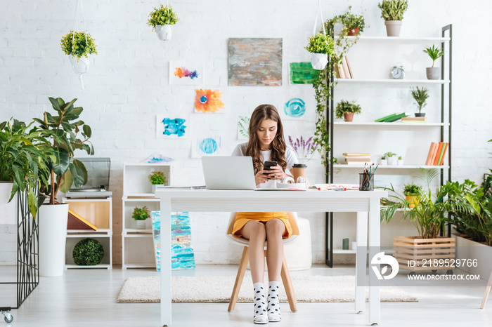 pretty young woman using smartphone while sitting in spacious room with green plants and paintings o