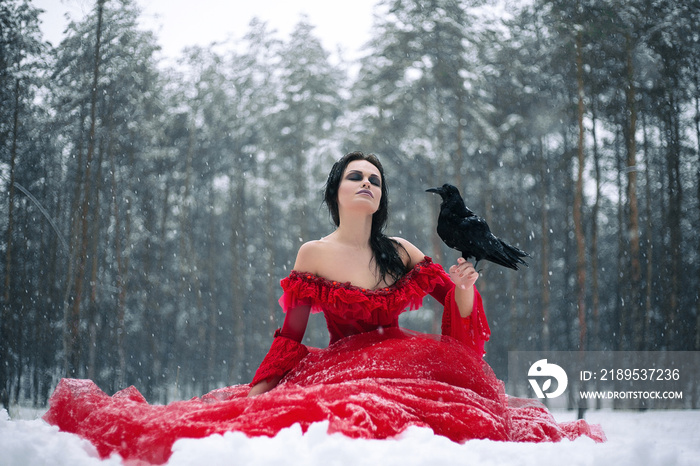 Woman witch in red dress with raven in her hand sits on snow in