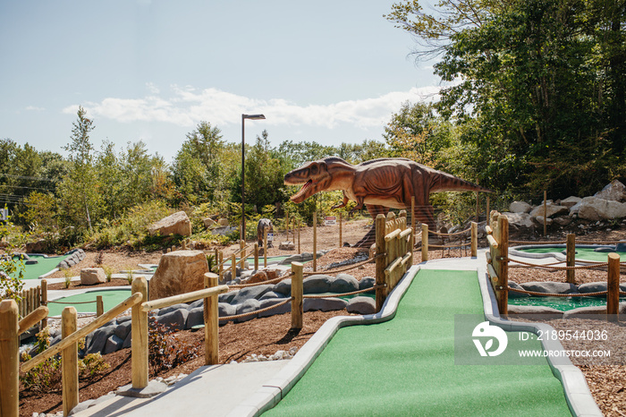 Green bridge leading to dinosaur sculpture and trees in miniature golf course against sky