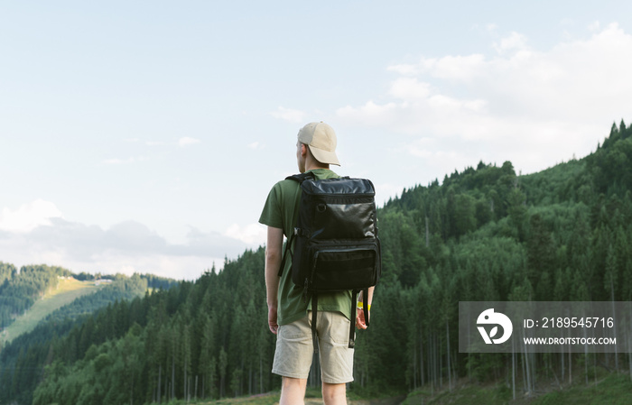Back view of young male traveler wearing cap with backpack standing alone in summer woods and lookin
