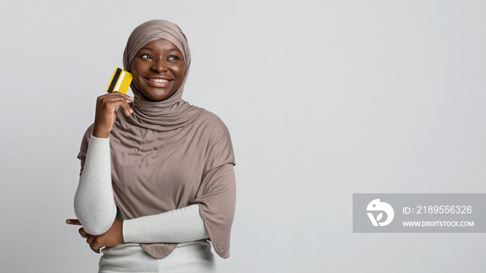 Pensive african muslim woman in hijab holding credit card and looking away