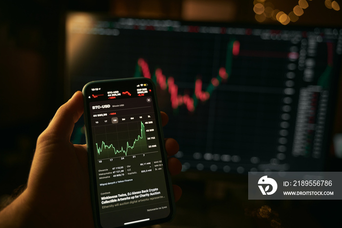 Hand holding a modern smartphone with the stocks market app and big screen blurred on the background