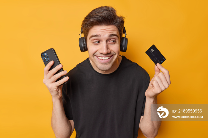 Photo of happy brunet man poses with credit card and smartphone orders in app pays online listens mu