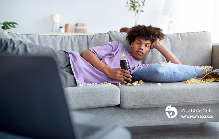 无聊的青少年躺在沙发上，手里拿着零散的零食，在家里用笔记本电脑看电影