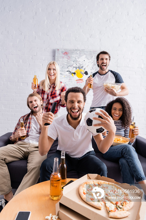 cheerful african american man holding soccer ball while watching football competition with excited m