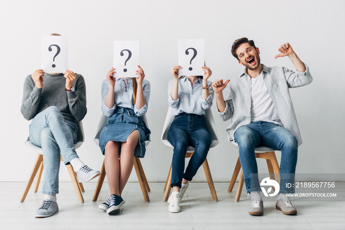 Cheerful man showing thumbs up near candidates with question marks on cards in office