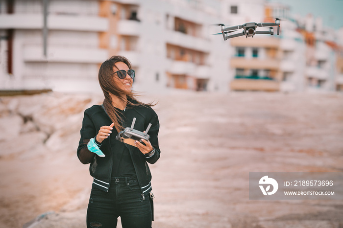 Pretty modern smiling woman pilot in dark stylish clothes flying drone with strong wind in city. Fly