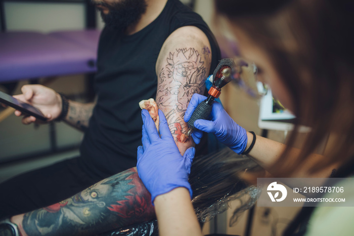 tattooist woman makes a tattoo in her studio to a young man while consulting his smartphone