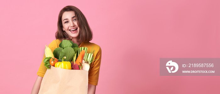 Happy woman holding paper bag full of fresh vegetable groceries isolated on pink copy space backgrou