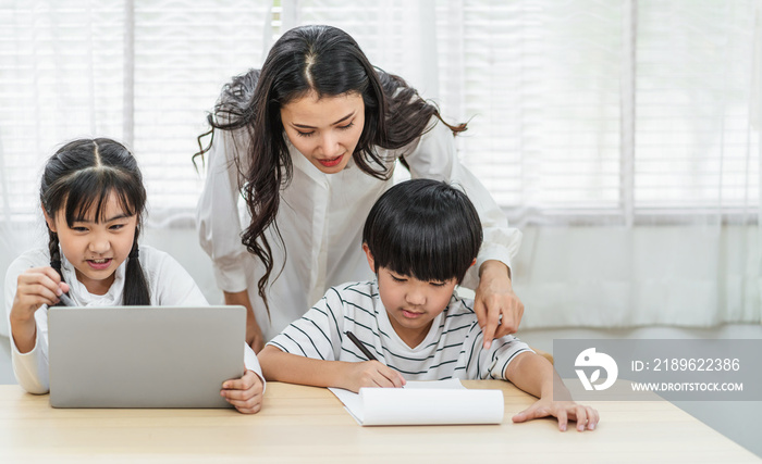 Young Asian Single Mother teaching or helping daughter and son doing home work with technology lapto