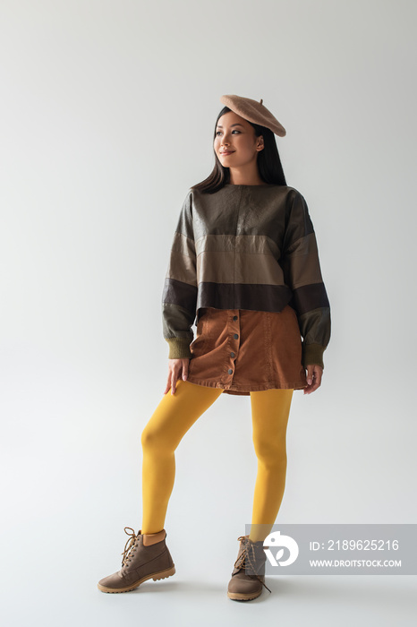 full length view of smiling asian woman in fashionable clothes and yellow tights looking away on gre