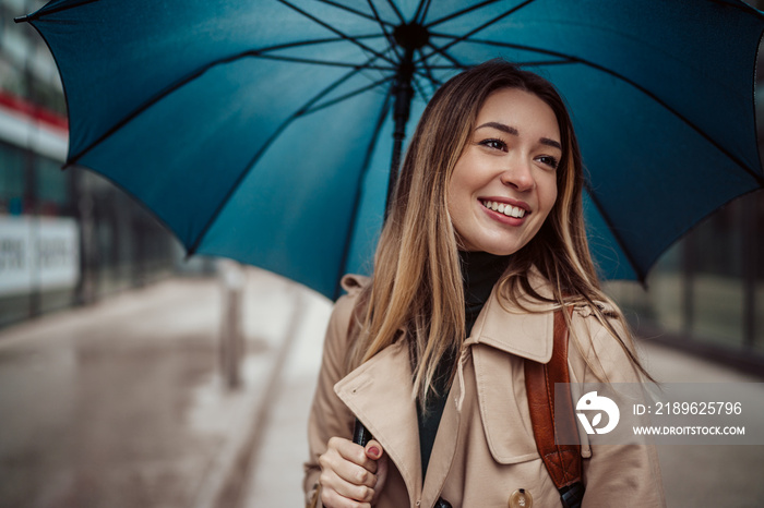 A beautiful smiling young woman walking through the city with an umbrella.