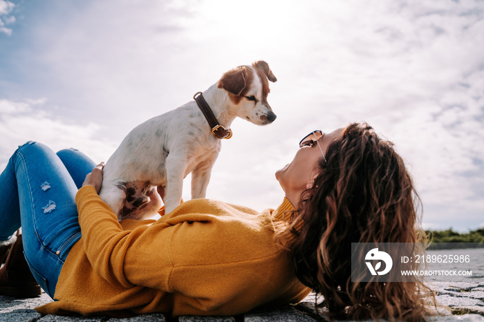 A beautiful little dog sitting on its owner looking at her face. The woman is lying down in the park