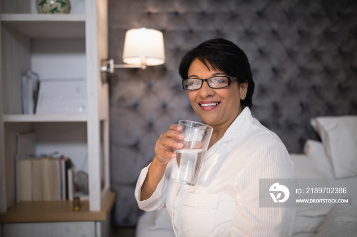 Smiling mature woman holding water glass in bedroom at home