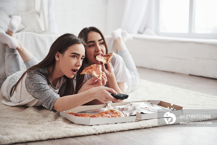 Leisure time. Sisters eating pizza when watching TV while lying on the floor of beautiful bedroom at