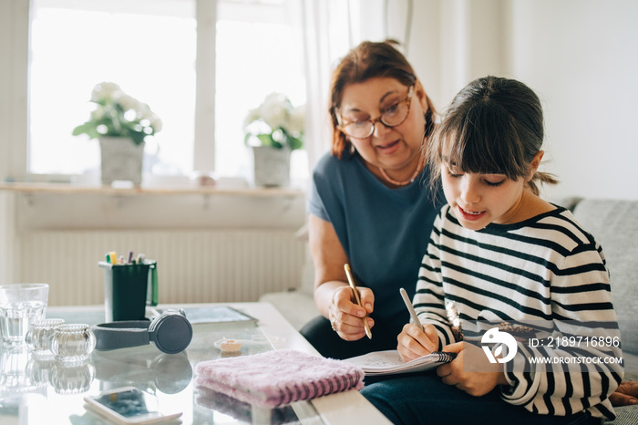 Grandmother looking at granddaughter doing homework on sofa at home