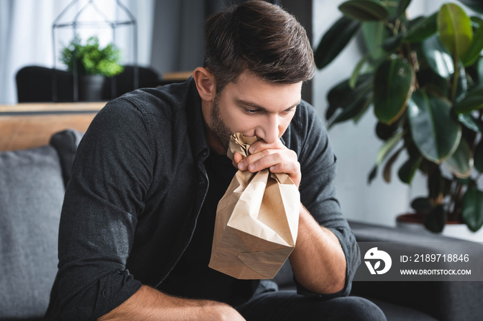 handsome man with panic attack breathing in paper bag in apartment