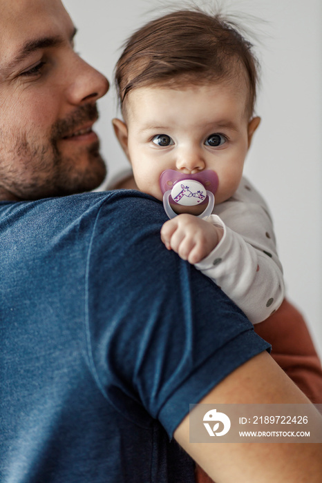 一个带着奶嘴的女婴和她的爸爸一起看着相机的肖像。