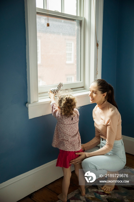 Smiling mother crouching by daughter playing with toy on window sill at home