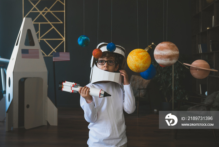 可爱的小男孩戴着纸板宇航员头盔，驾驶玩具火箭穿越行星，纸板太空舱