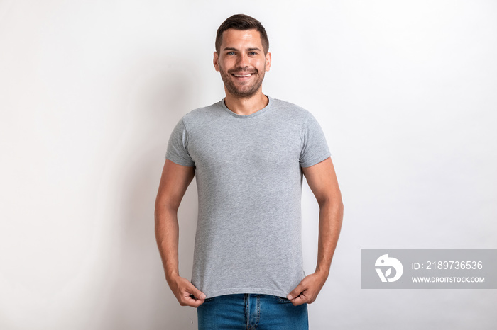 Funny man wearing in grey t-shirt standing pulling down a t-shirt smiling and looking at the camera.