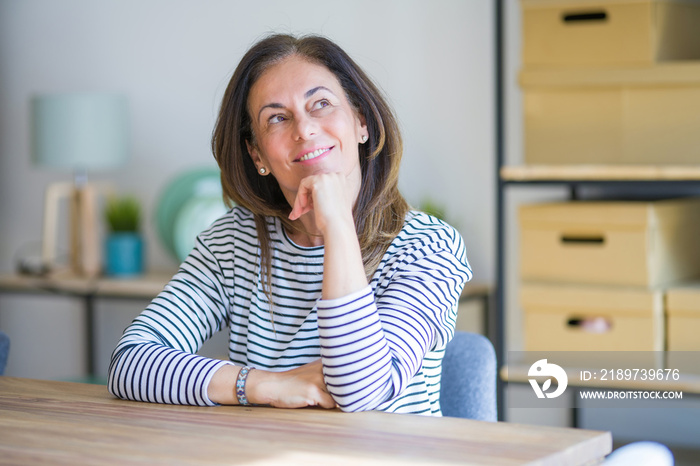 Middle age senior woman sitting at the table at home with hand on chin thinking about question, pens